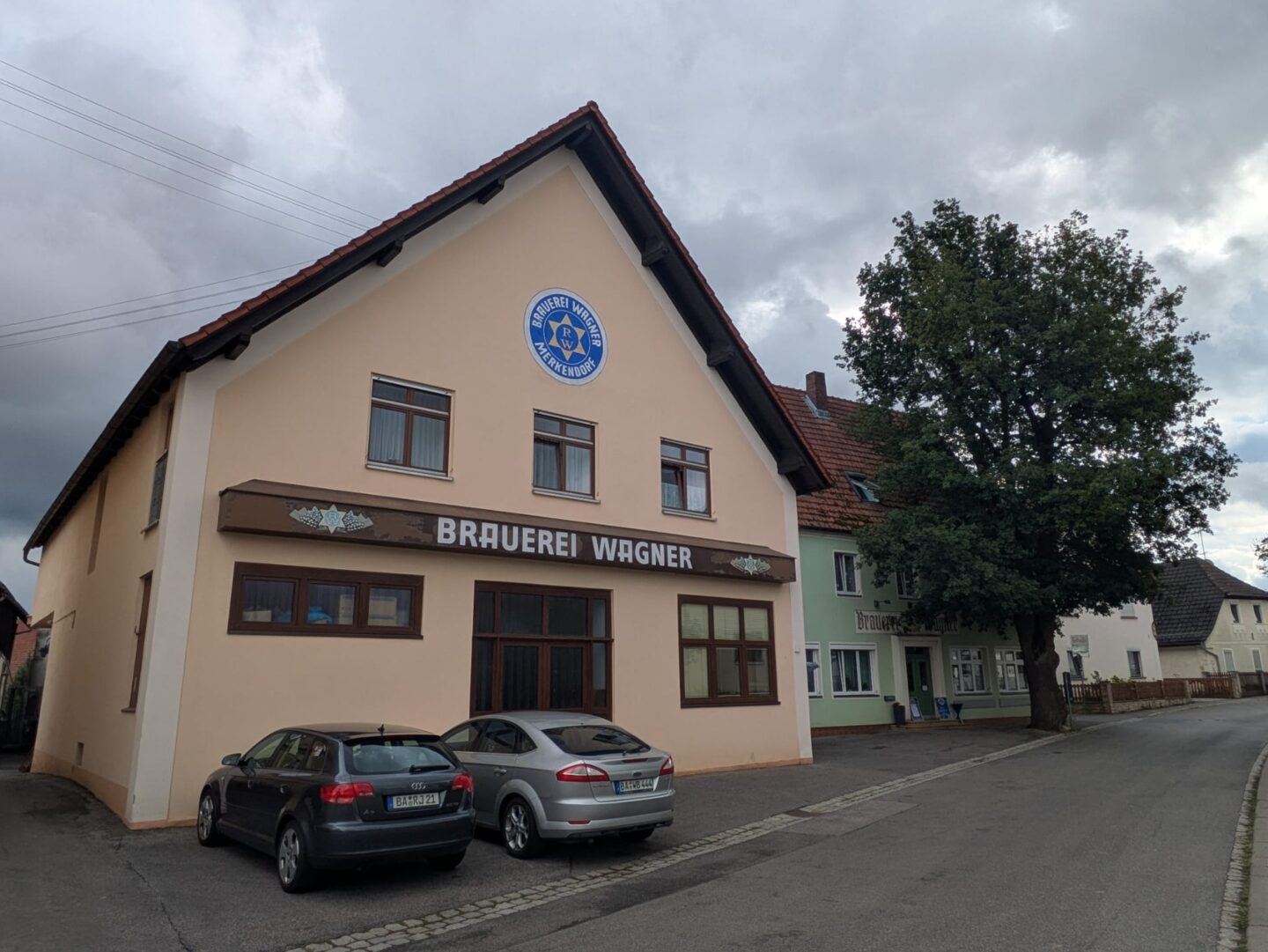 Biergarten der Brauerei Wagner: Schattenspende Bäume, blaugelber Brauereistern und zahlreiche Wanderer in Merkendorf